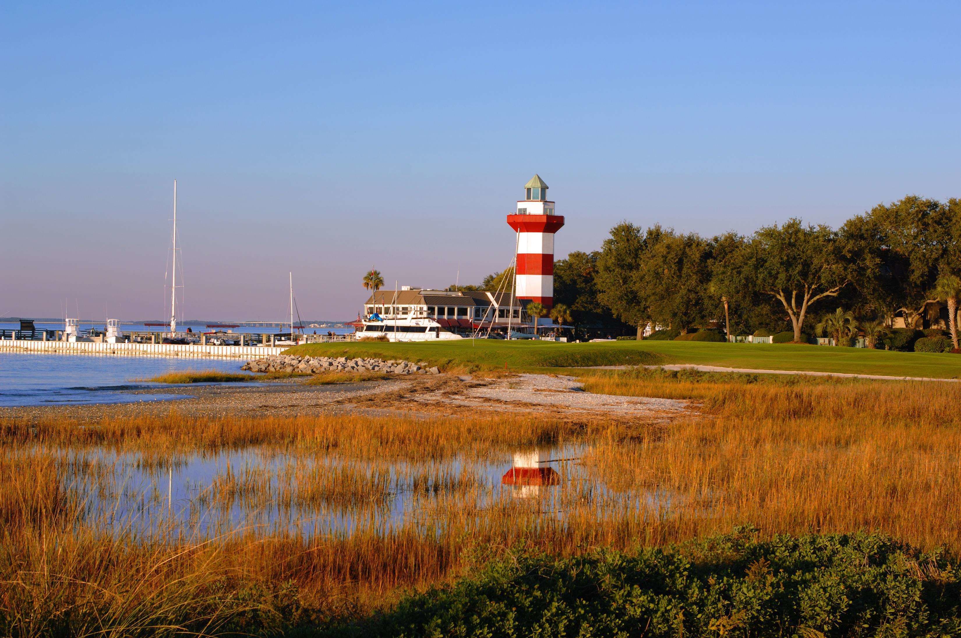 A lighthouse with a red and white striped tower, a dock with boats, and a grassy area with a small pond.