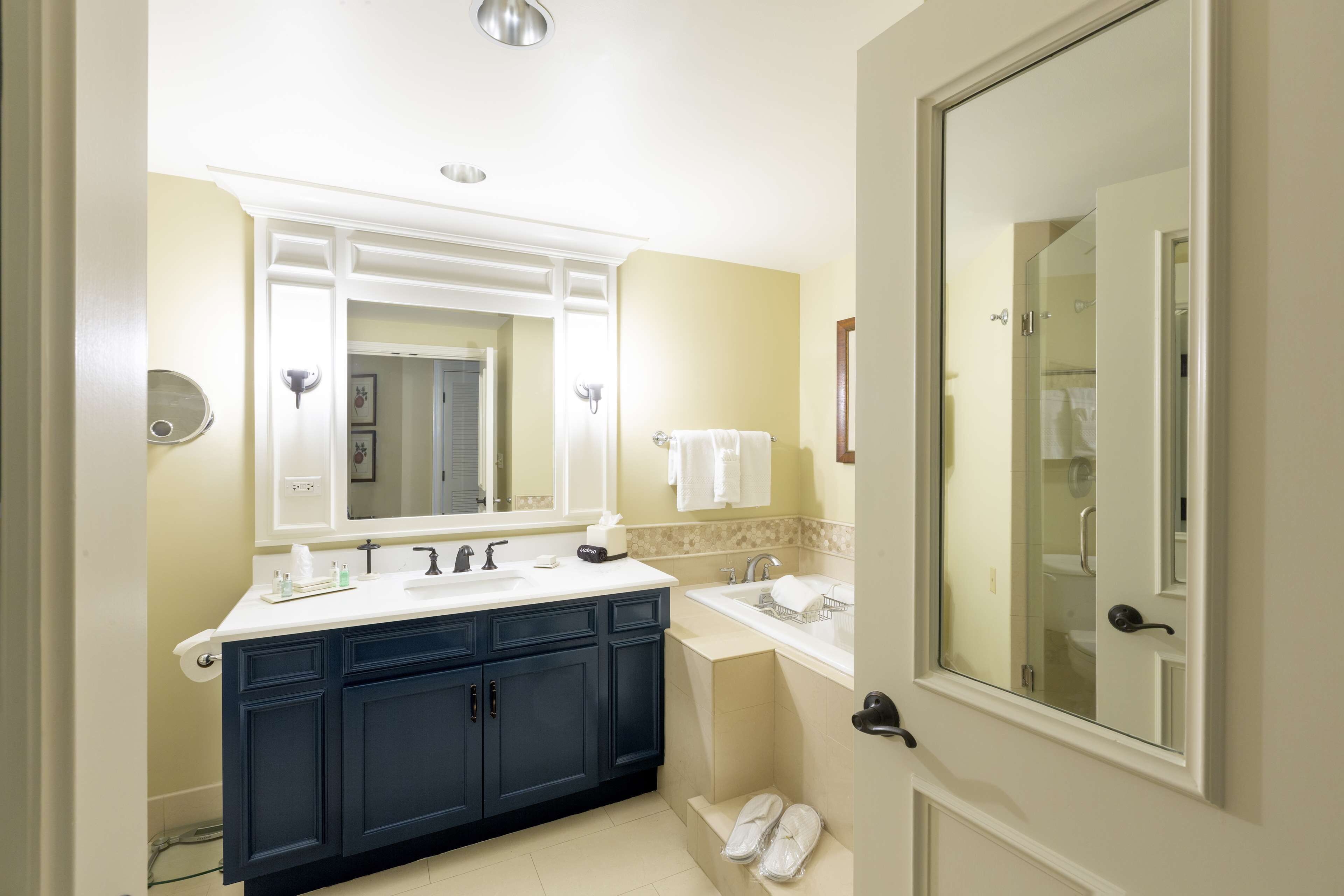 A bathroom with a double sink, dark blue cabinets, a large mirror, and a glass shower door.