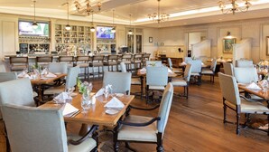 A restaurant with multiple dining tables and chairs, a bar area with a television, and a variety of bottles and glasses on shelves. - The Inn & Club at Harbour Town (Hilton Head Island)