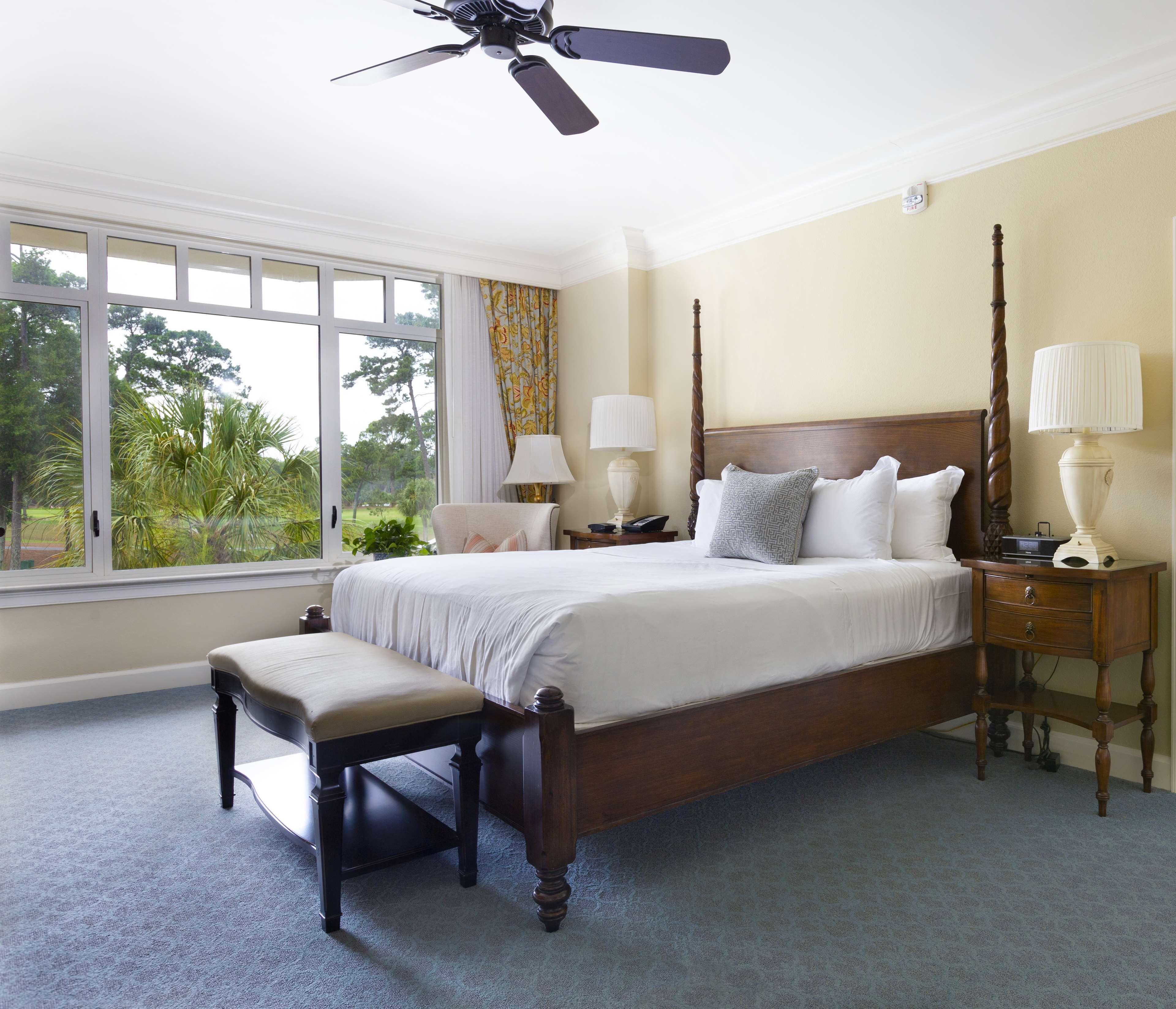 A bedroom with a four-poster bed, a bench, two bedside tables with lamps, and a ceiling fan.