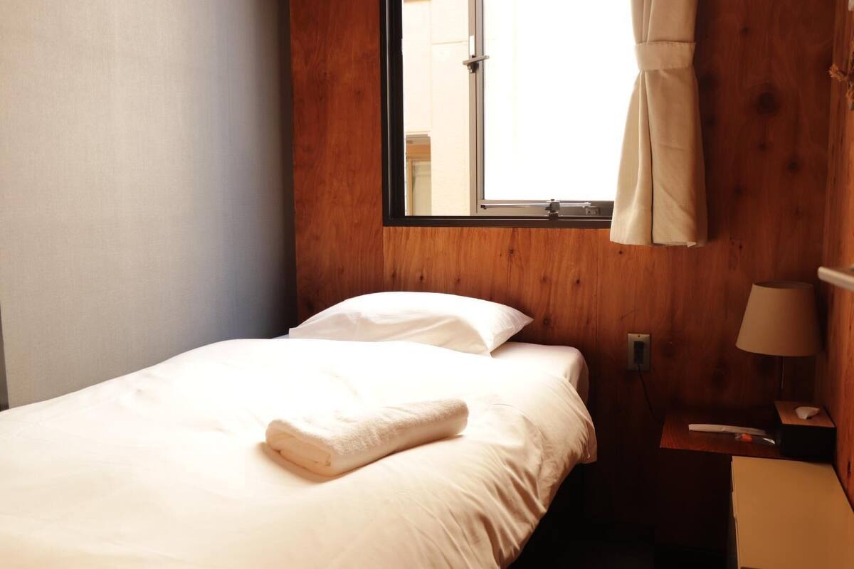 Una cama individual con ropa blanca, una ventana con cortinas, una mesita de noche con lámpara y una pared de madera.