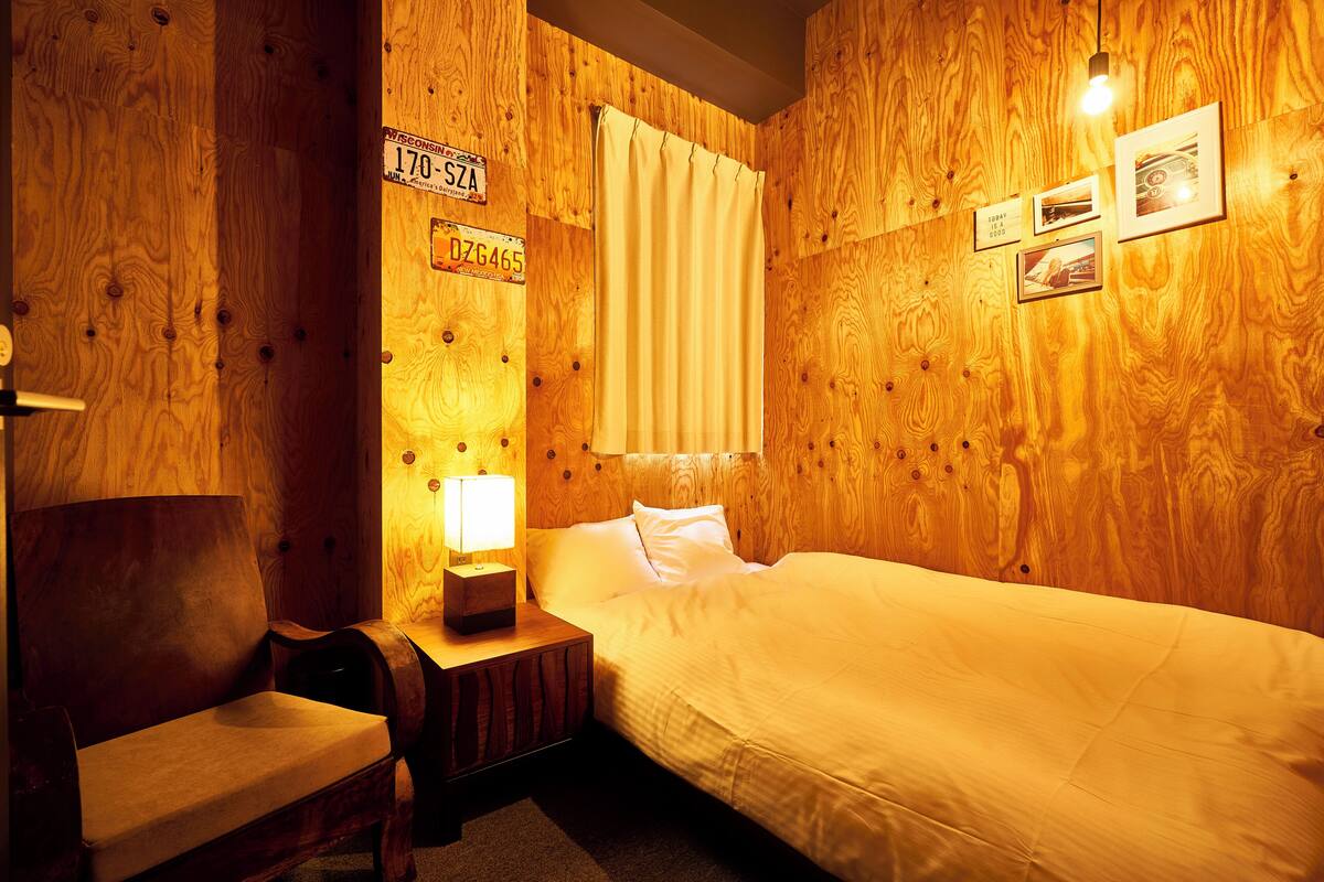 Una habitación con paneles de madera, una cama, una mesita de noche con lámpara, una silla y una ventana con cortinas.