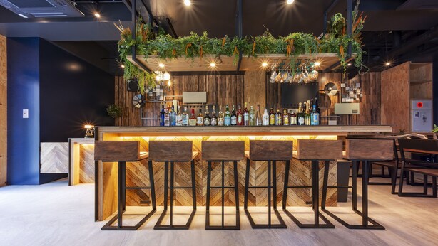 A bar area with a wooden bar counter, multiple stools, and a variety of bottles. - FOCUS Kuramae - Hostel (Tokyo)