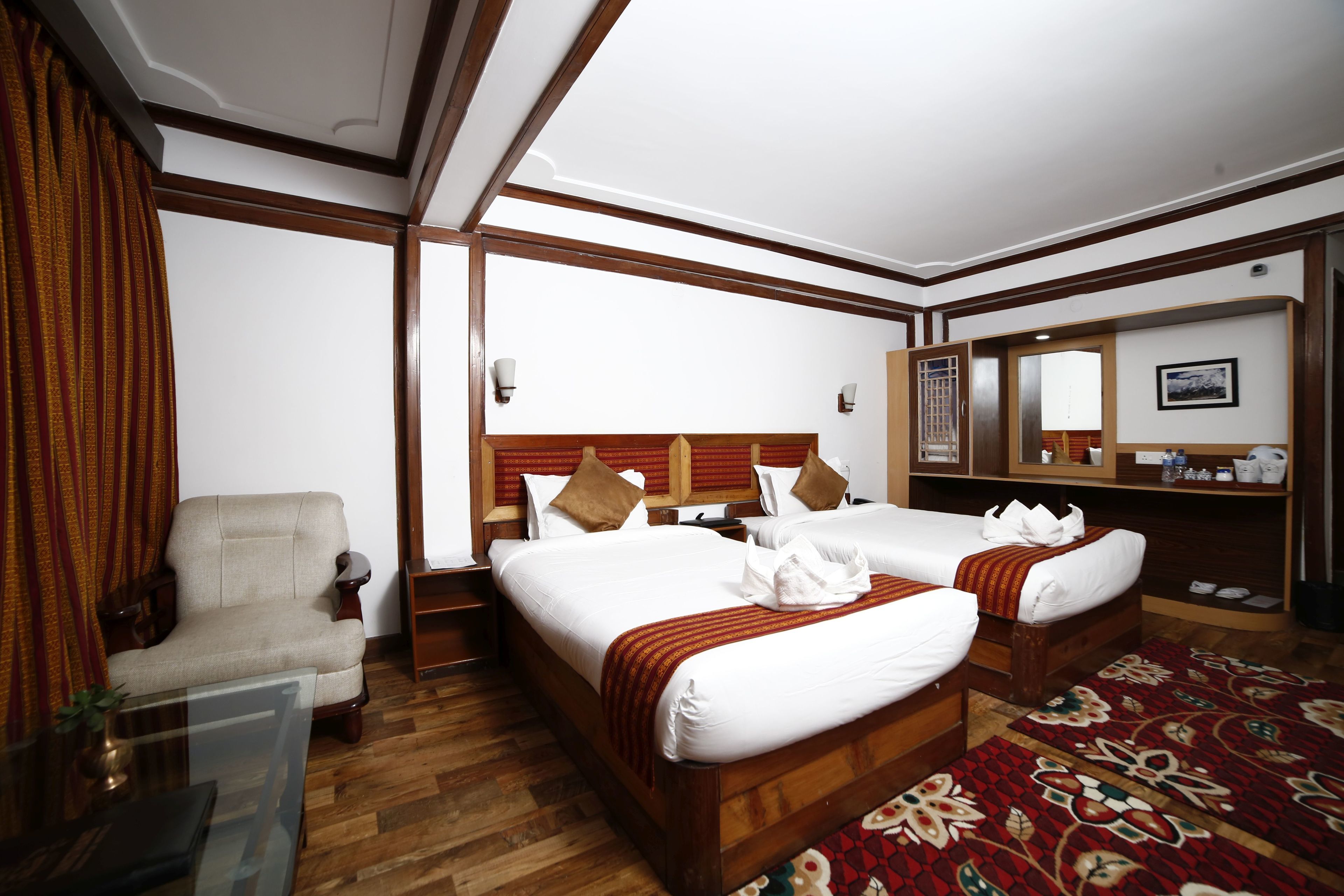 A hotel room with two beds, a wooden headboard, a chair, a glass table, and a red patterned rug.