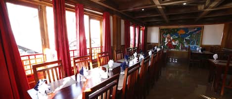 A restaurant with wooden tables and chairs, large windows with red curtains, and a mural on the wall.