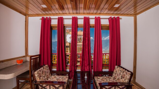 A room with wooden ceiling, red curtains, two wooden chairs, a small table, and a view of a building through the window. - Buddha Retreat (Namchi)