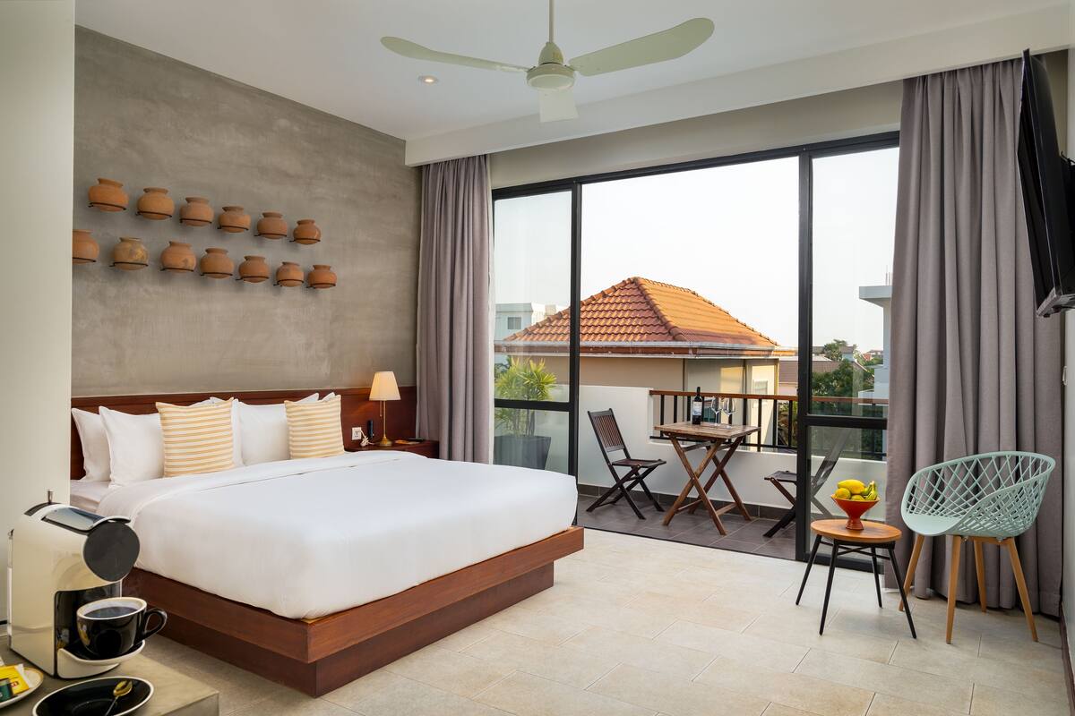 A hotel room with a large bed, a balcony with a table and chairs, and a view of a building with a red roof.