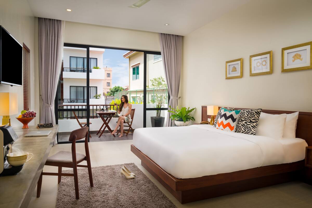 A modern hotel room with a large bed, a desk with a chair, a TV, and a balcony with a view of the city.