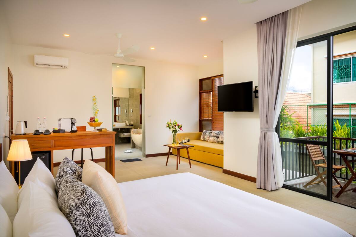 A hotel room with a bed, a desk with a lamp, a television, and a balcony with a table and chairs.