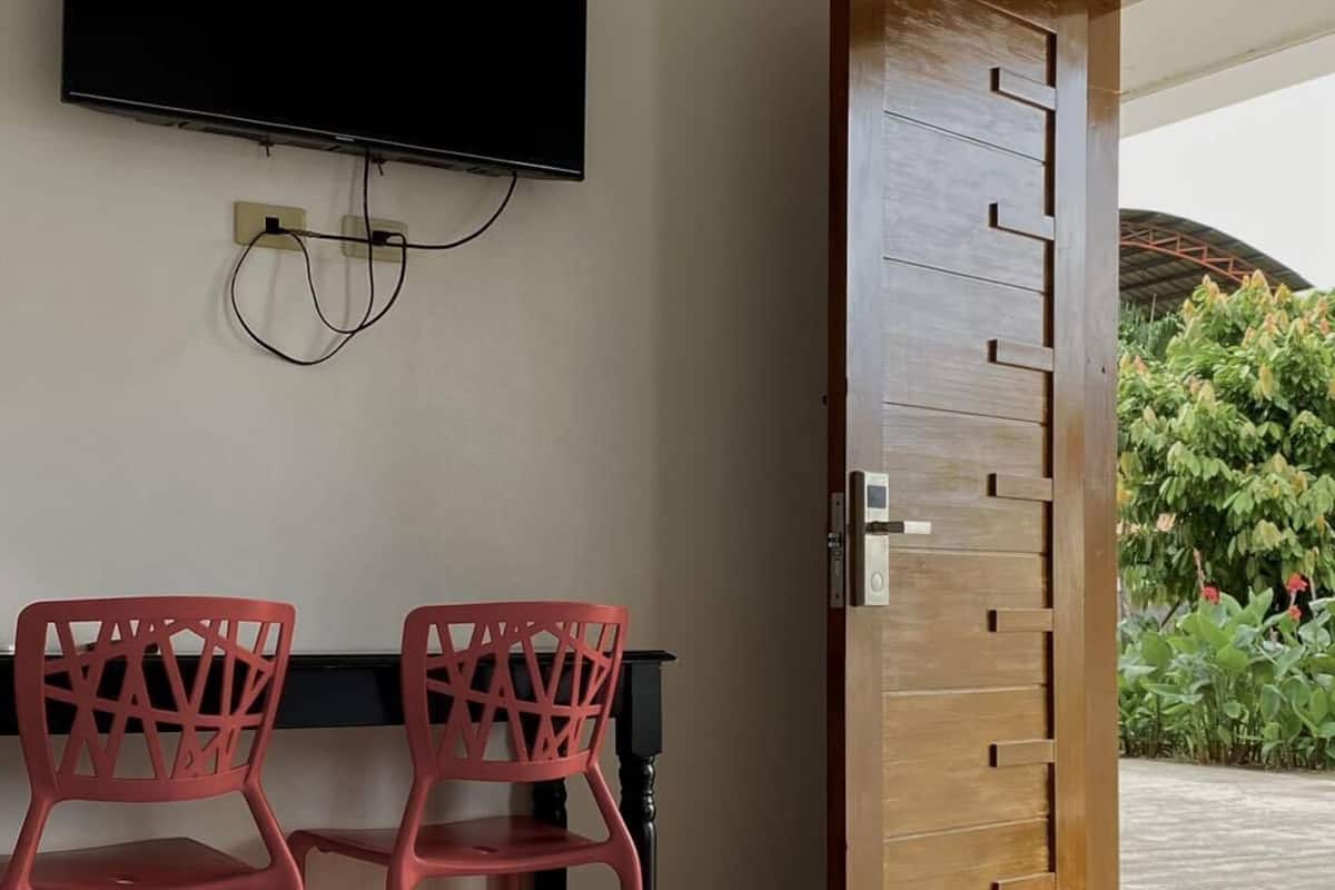 Un dormitorio con una cama, dos sillas rojas, un televisor montado en la pared y una puerta de madera que da a un espacio exterior.
