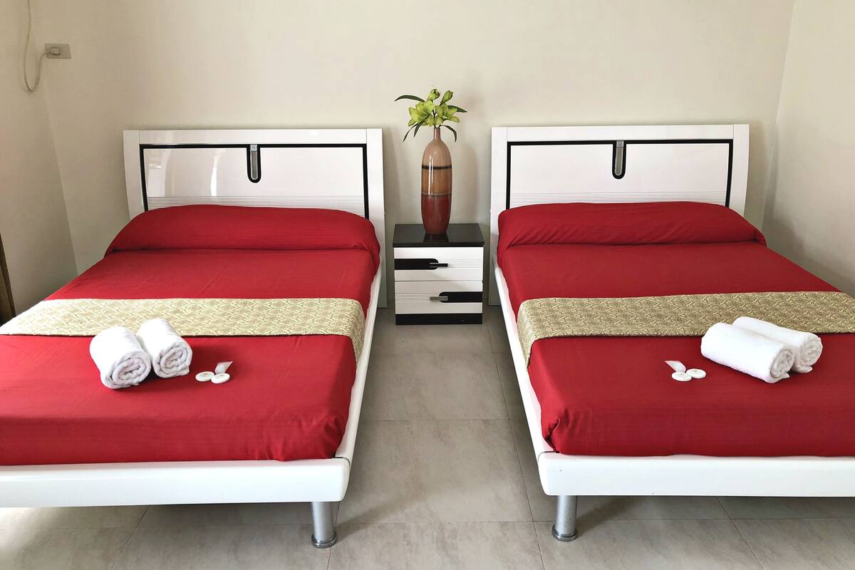 Habitación de hotel con dos camas, cada una con ropa de cama roja y una colcha estampada color beige. Hay dos mesitas de noche blancas con cajones, un jarrón con una planta y una mesita pequeña con una vela.