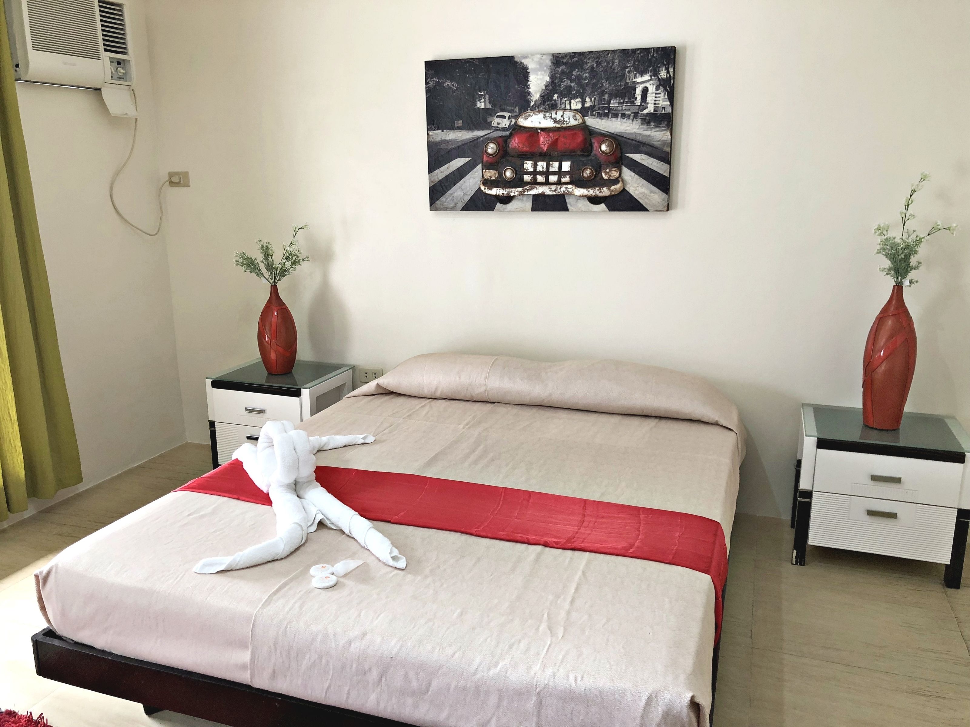A neatly made bed with a red accent, two bedside tables with vases, and a wall-mounted painting of a car.