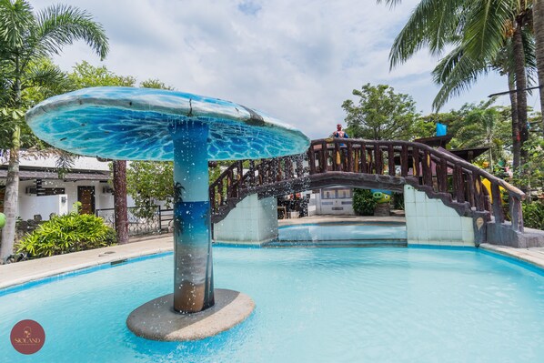 Una piscina con una fuente central, rodeada de palmeras y un puente de madera.
