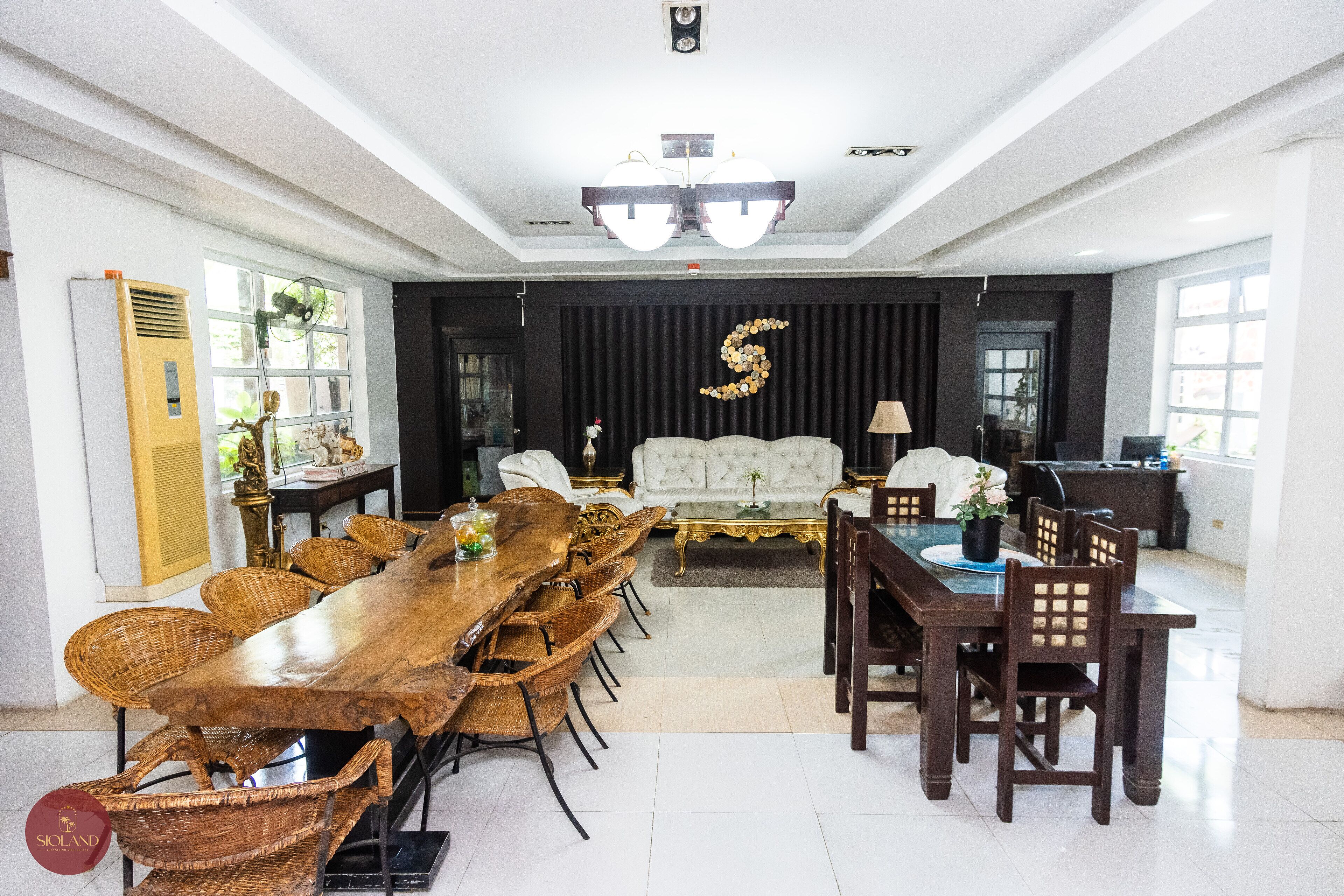 A spacious room with a long wooden dining table surrounded by wicker chairs, a white sofa, and a dark curtain backdrop.
