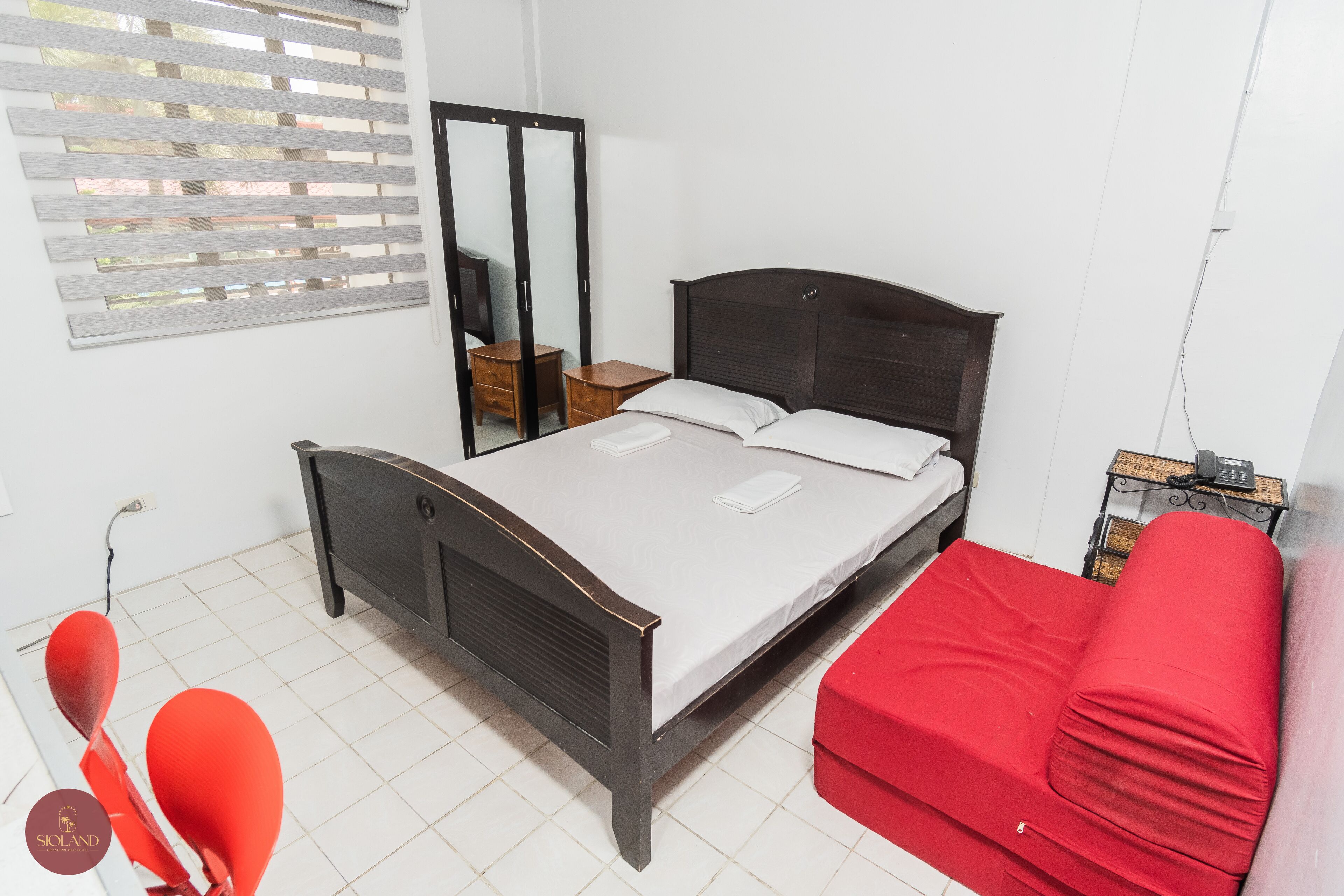 A bedroom with a bed, a red sofa, a wooden wardrobe, and a mirror.