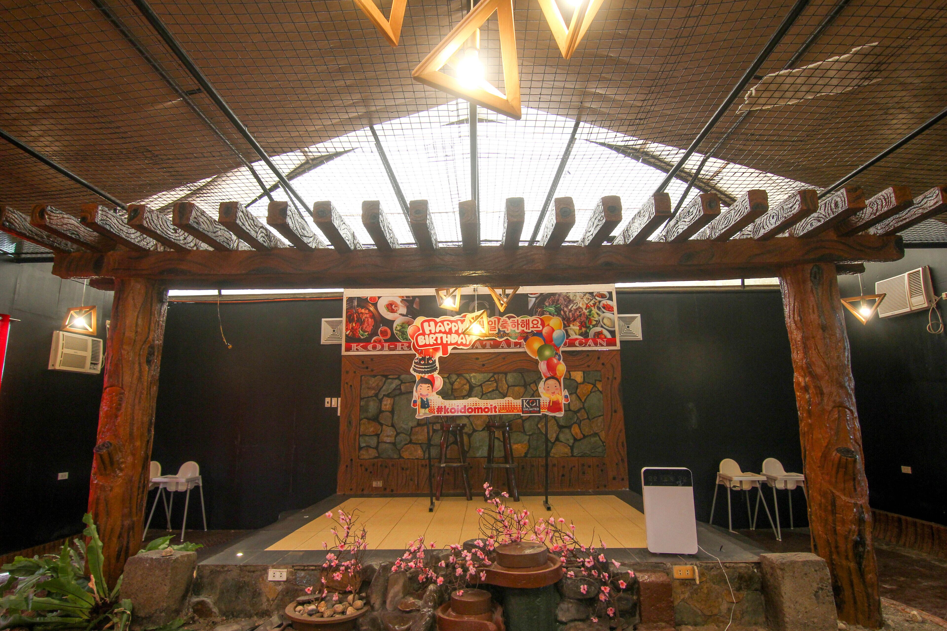A covered outdoor area with a stone wall backdrop, a wooden pergola, and a birthday celebration setup including a banner and balloons.