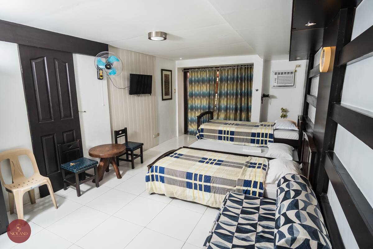 A bedroom with two beds, a chair, a table, a fan, and a TV.