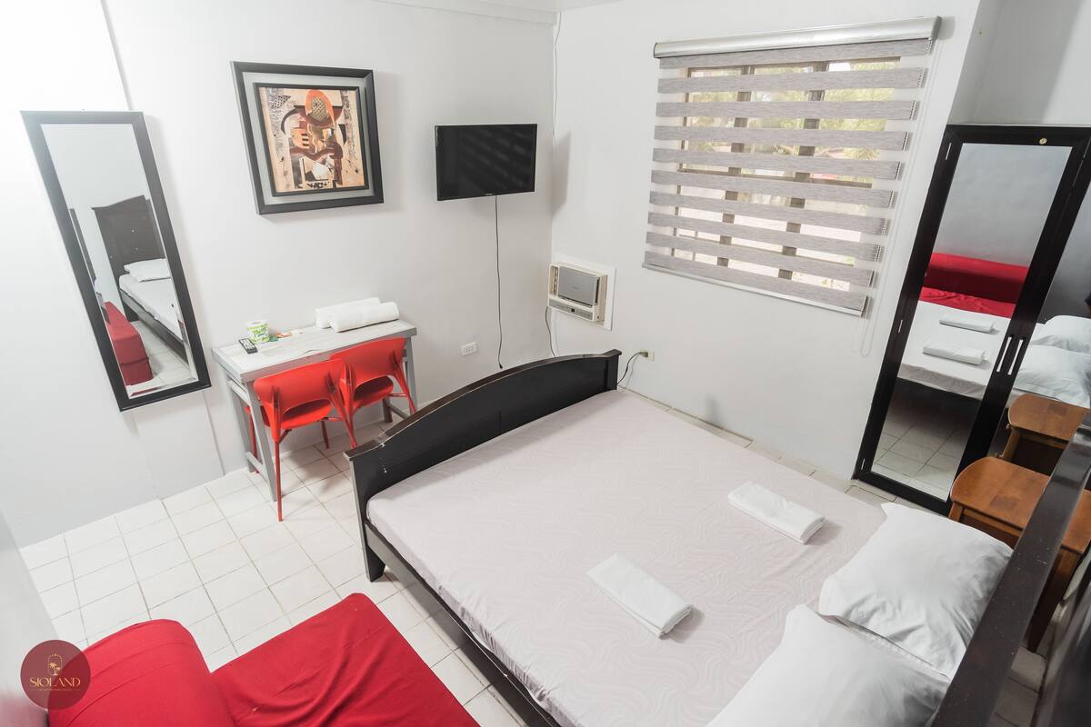 A hotel room with a bed, a desk with a chair, a TV, a window with blinds, and a mirror.