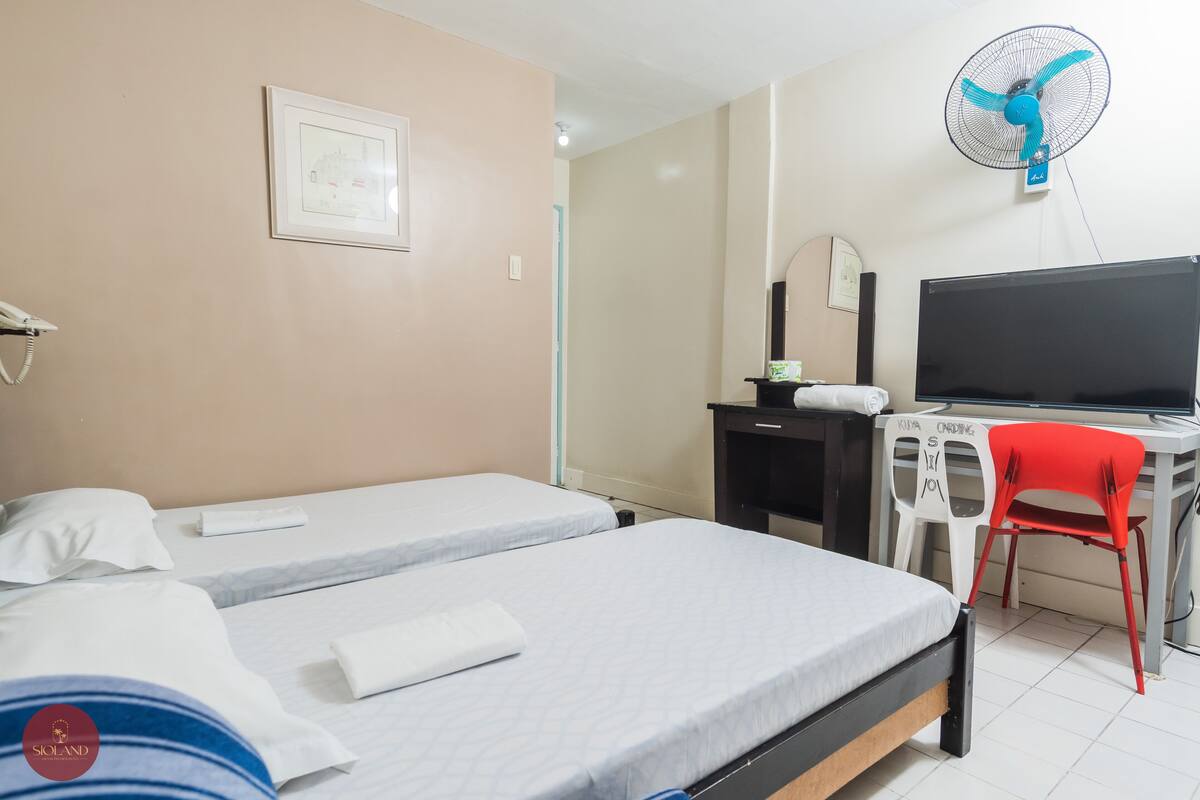 A hotel room with two beds, a desk with a computer, a red chair, a wall-mounted fan, and a framed picture on the wall.