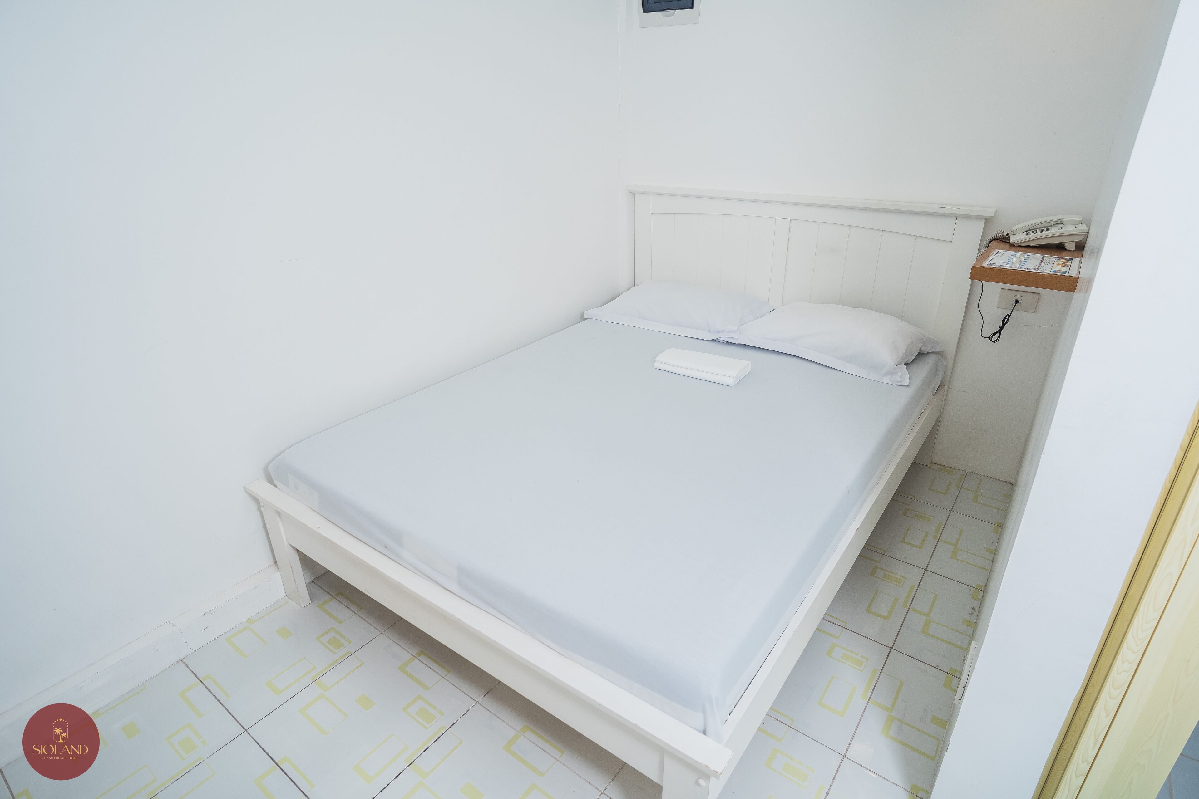 Ein kleines, weißes Zimmer mit einem Einzelbett, schlichtem Design, einem Nachttischtelefon und einer kleinen Ablagefläche.