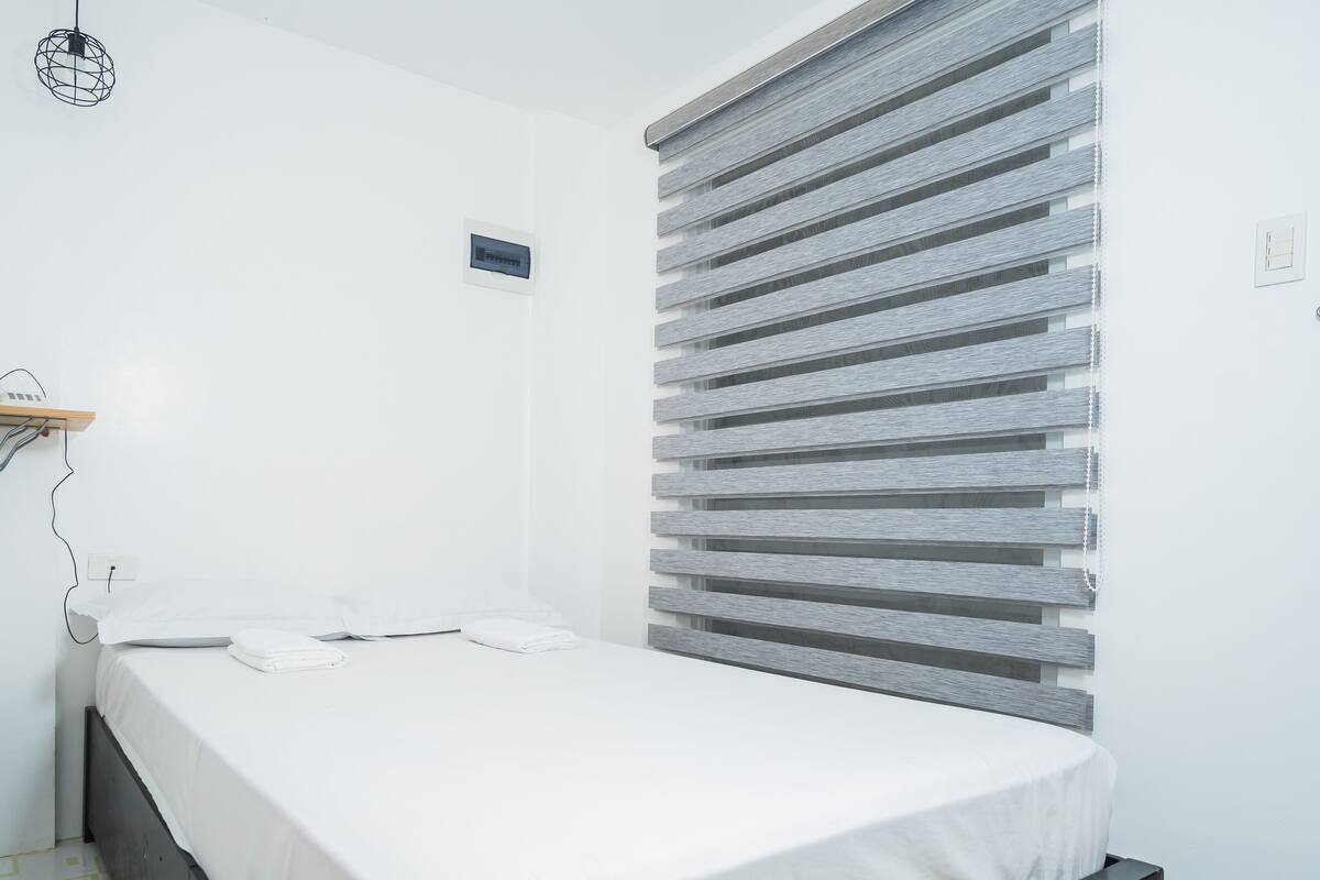 A single bed with a white mattress and pillows, a bedside table, a wall-mounted heater, and a window with horizontal blinds.