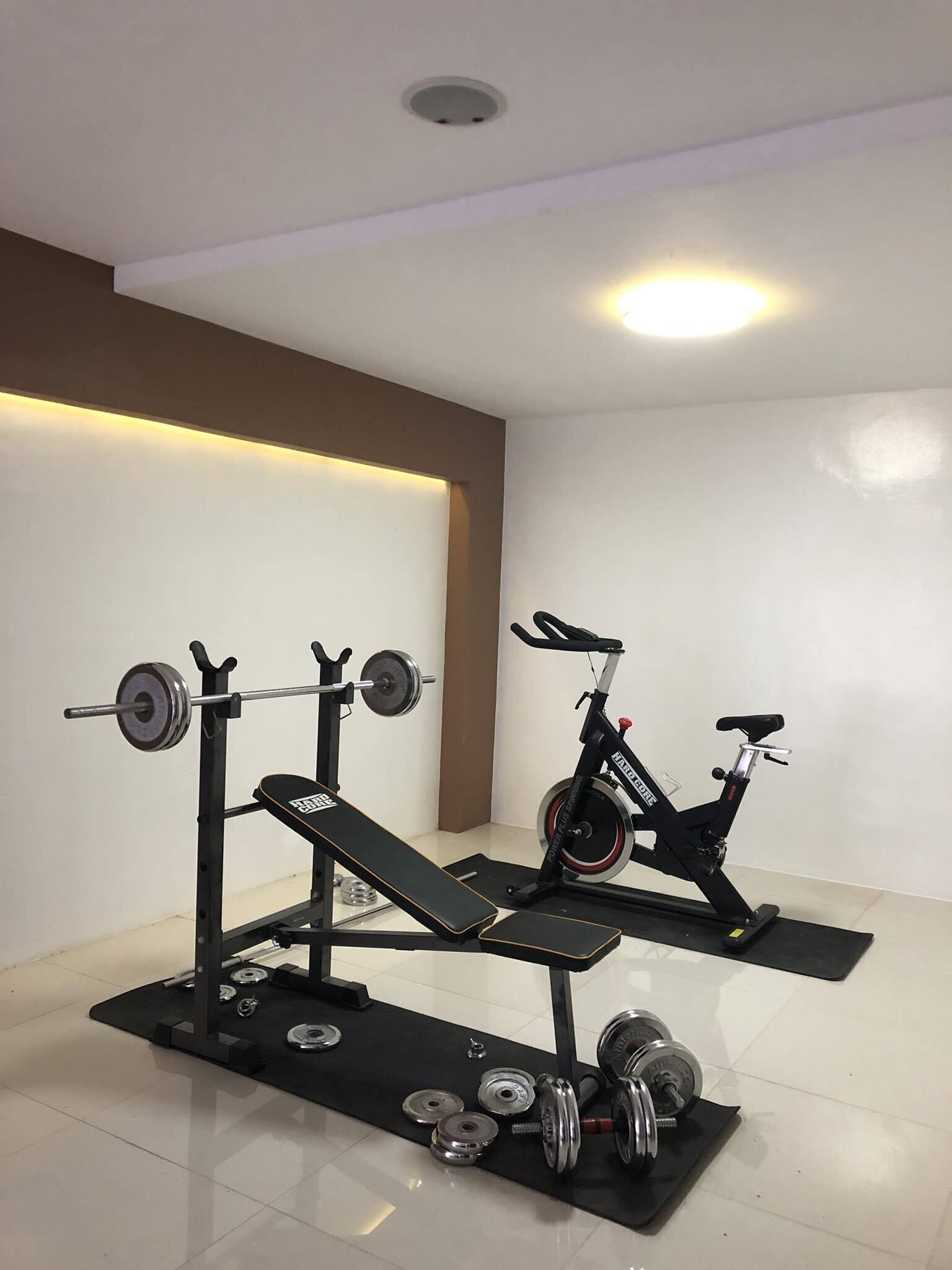 Une salle de sport à domicile équipée d’un banc de musculation, d’haltères, d’une barre et d’un vélo d’exercice.