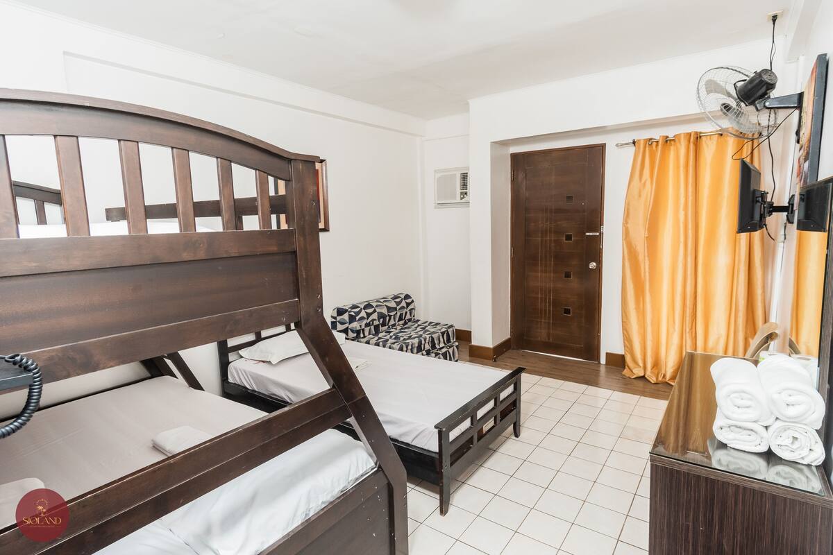 A hotel room with two wooden beds, a small sofa, a mirror, a fan, and a wooden dresser with towels.