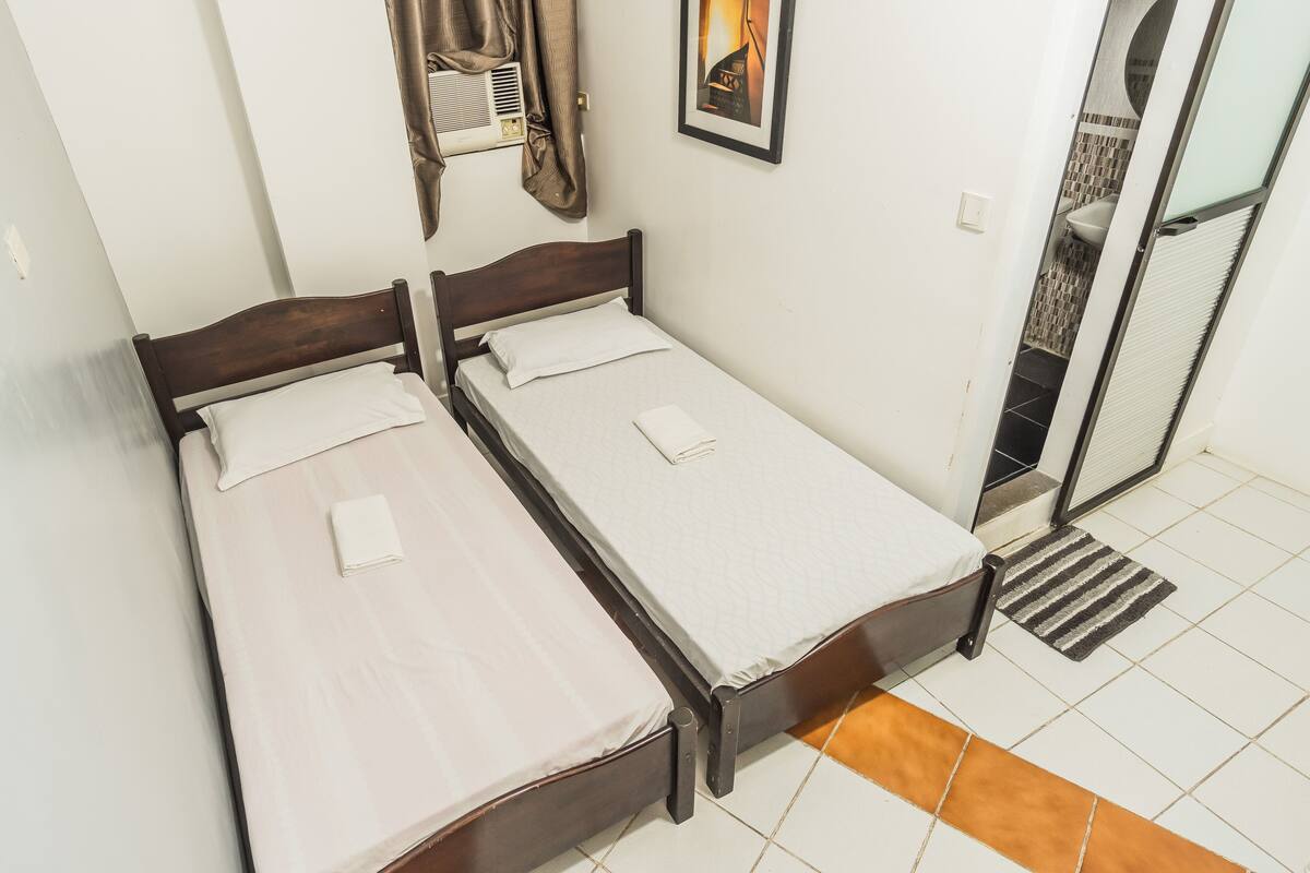 A double bed with a wooden frame, a white mattress, and a folded white towel. There is a framed picture on the wall and a window with curtains.