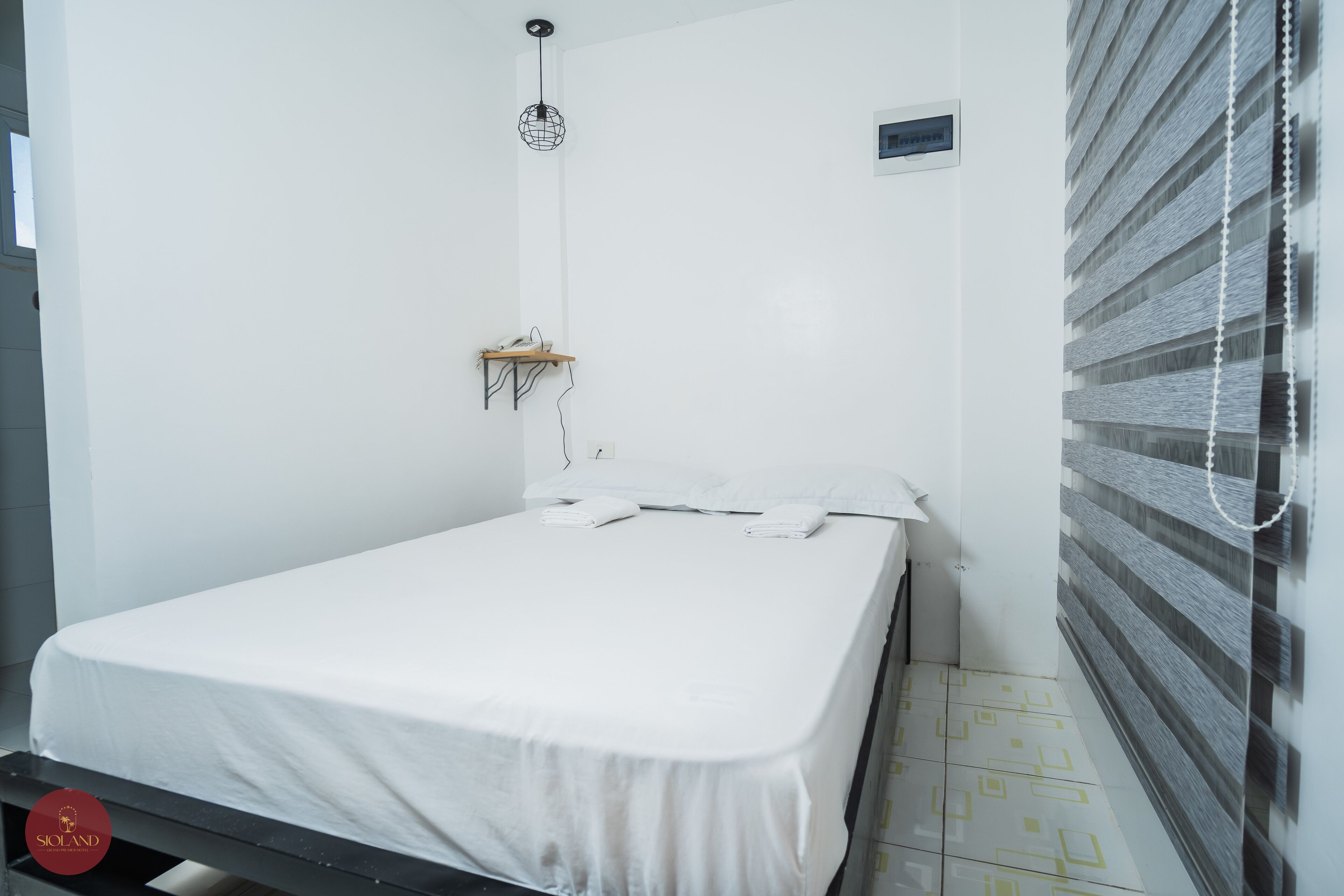 A hotel room with a bed, a small table, a wall-mounted shelf, and a hanging light fixture.