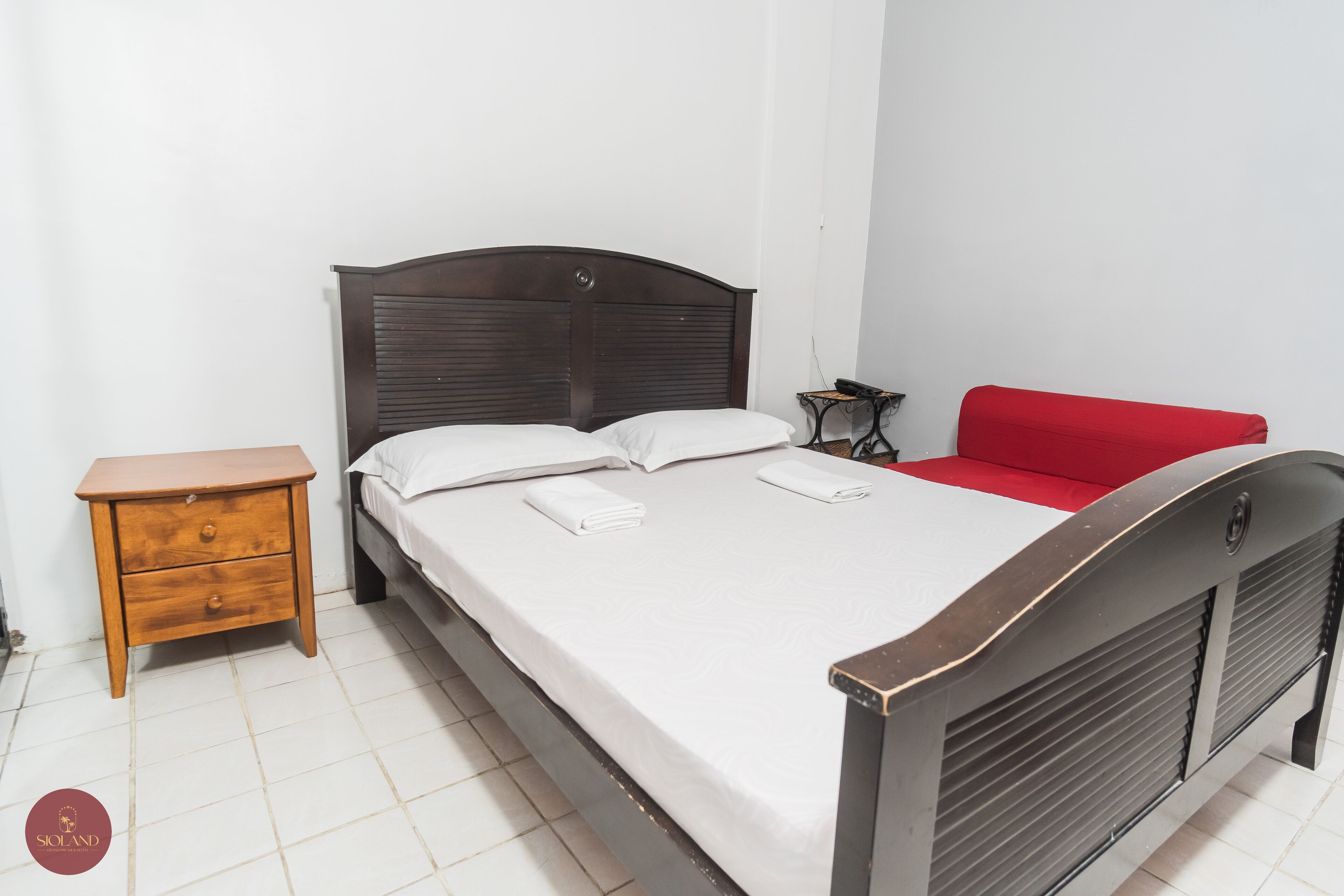 Una cama bien tendida con sábanas blancas, una mesita de noche de madera con cajones, un sofá rojo y una pequeña mesa con un teléfono.