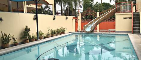 A swimming pool with a slide, surrounded by potted plants and a covered area with a metal roof.