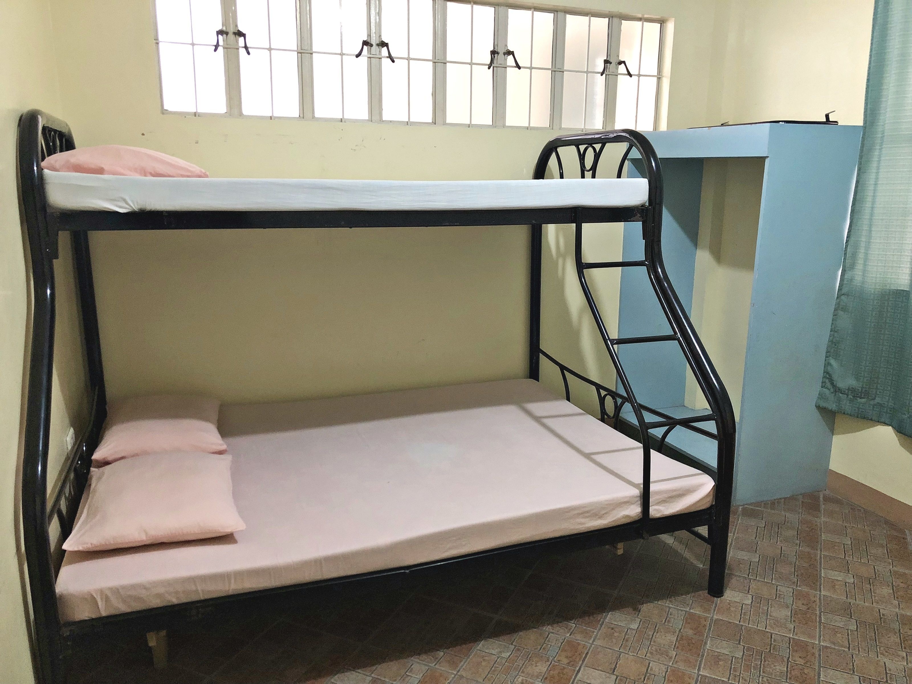 Una cama litera con colchón y almohadas, una ventana con barrotes y un armario azul.