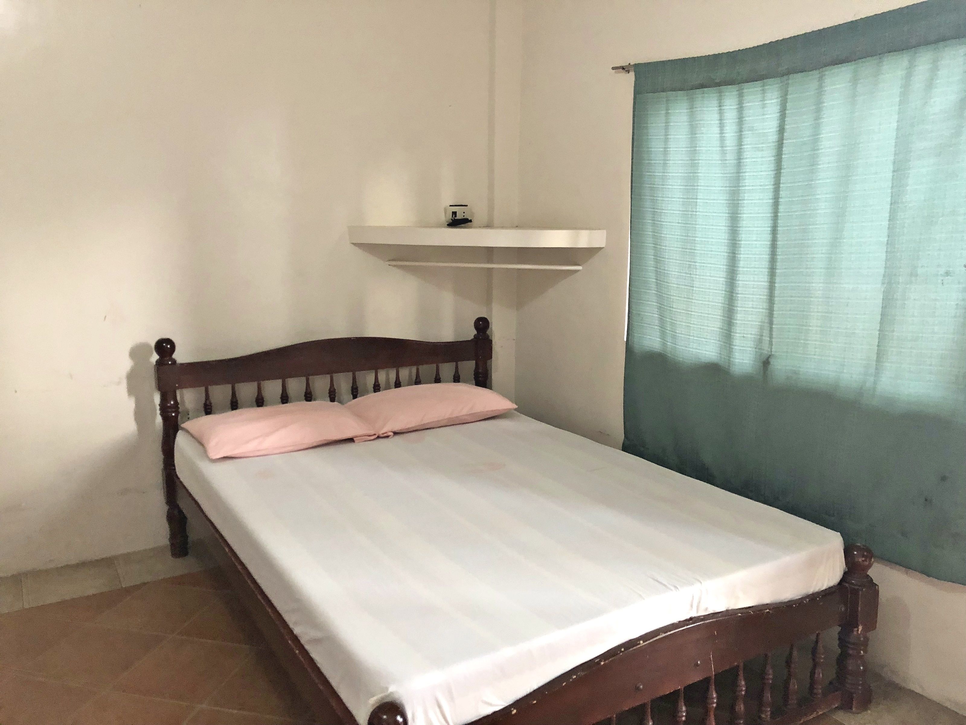 A single bed with a wooden frame and a white mattress, a small shelf above it, and a window with a green curtain.