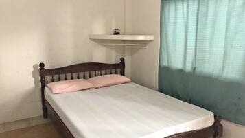 A single bed with a wooden frame and a white mattress, a small shelf above it, and a window with a green curtain.