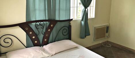Una cama con cabecera de madera, una ventana con cortinas y un aire acondicionado fijado a la pared.