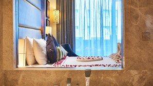 Une chambre d’hôtel avec une grande fenêtre, un lit avec des oreillers, une table de chevet avec une lampe et une salle de bain avec un miroir et un lavabo.