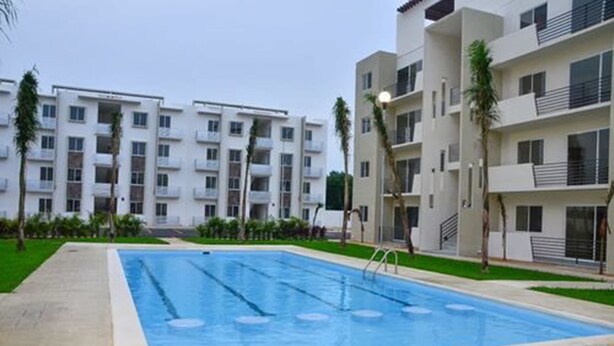 A swimming pool surrounded by modern apartment buildings with balconies and a well-maintained lawn. - Departamento vacacional (Playa del Carmen)