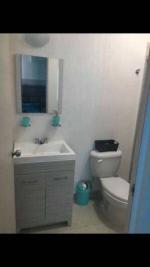 A small bathroom with a white sink, mirror, and toilet. There is a trash can and a small cabinet below the sink. - Departamento vacacional (Playa del Carmen)