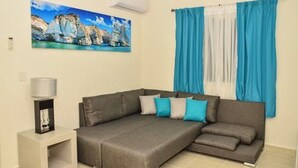 A living room with a gray sectional sofa, a framed picture on the wall, and a window with blue curtains. - Departamento vacacional (Playa del Carmen)