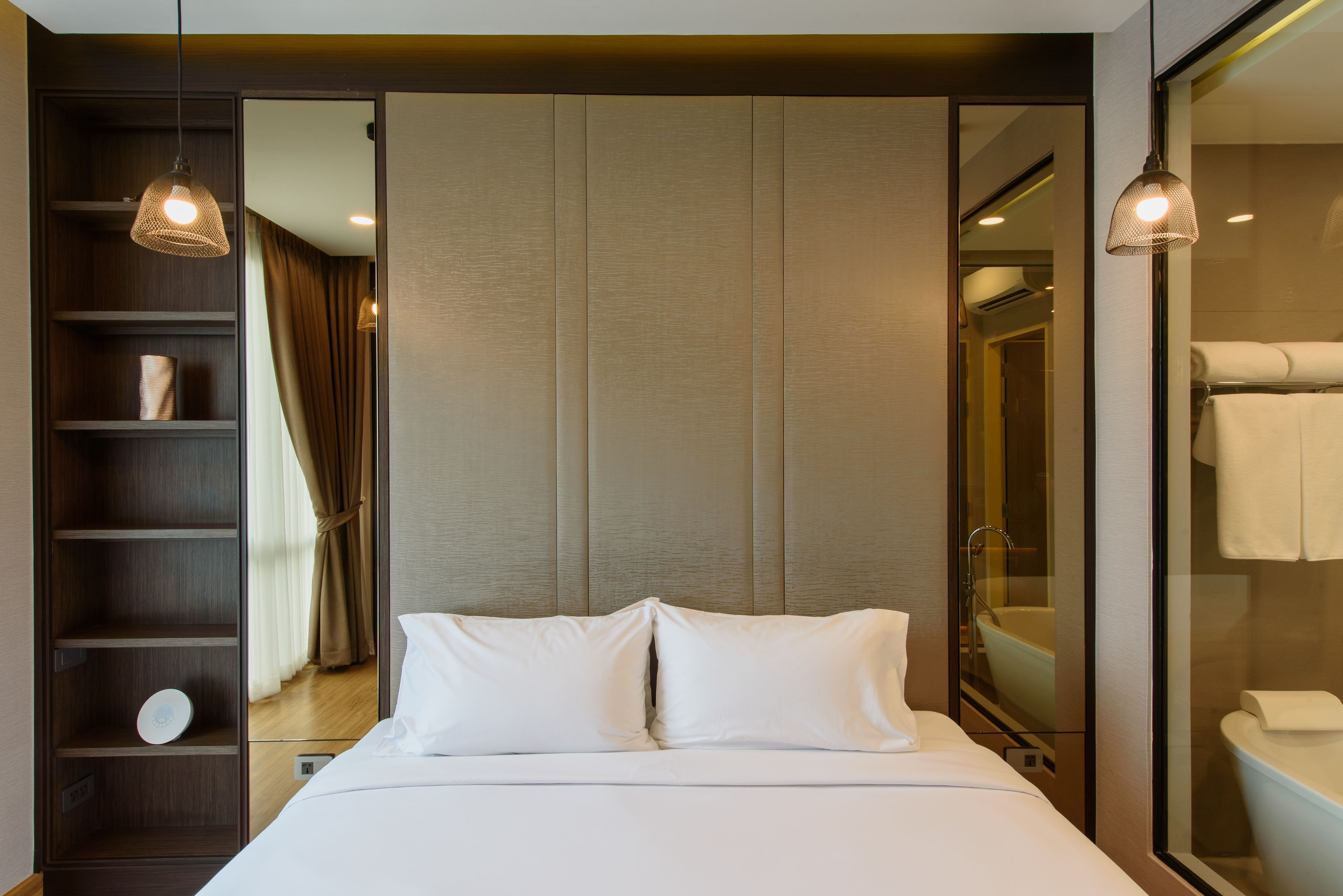 A modern hotel room with a large bed, a wardrobe, a mirror, and a bathroom visible through a glass partition.