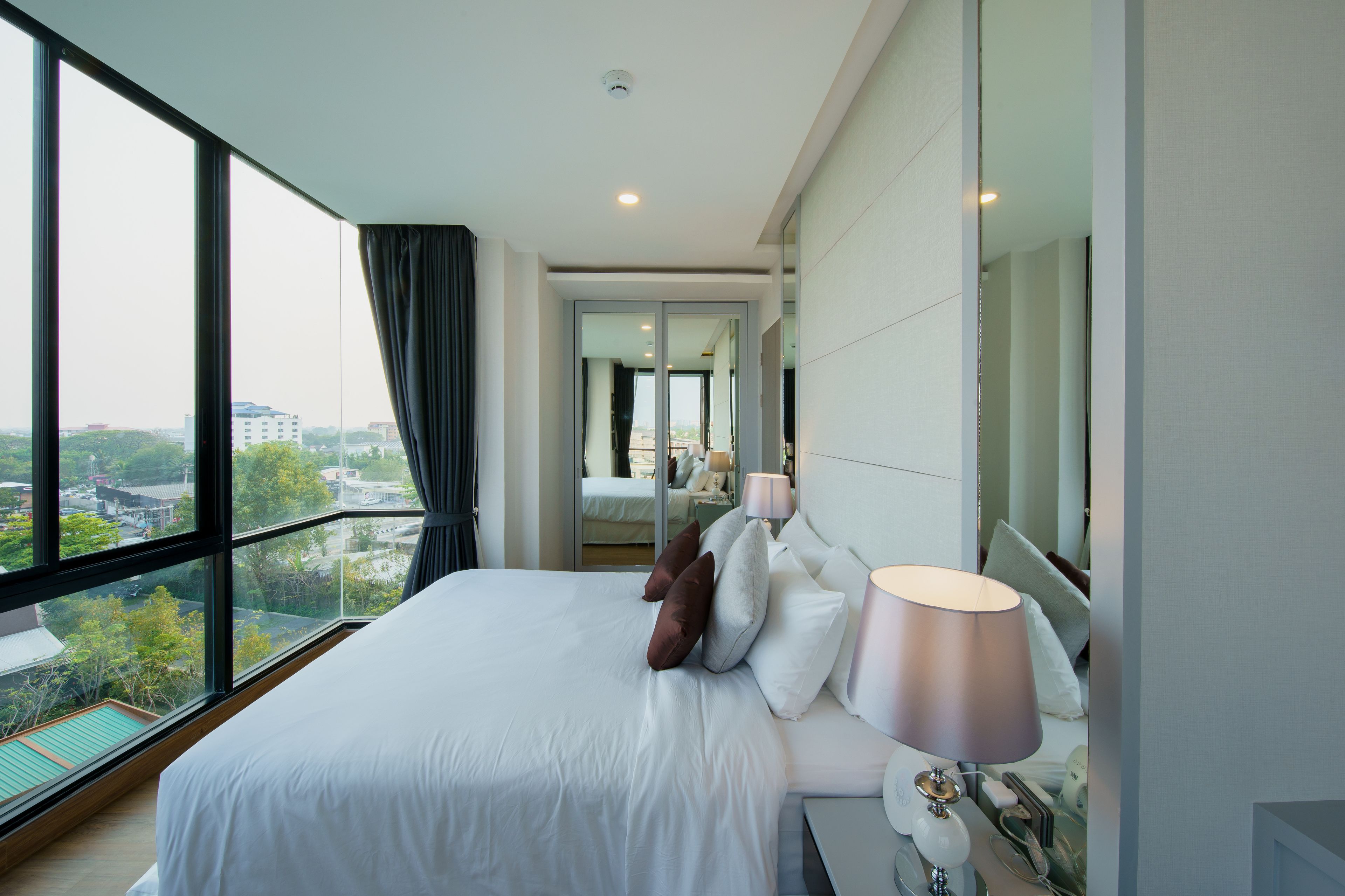 A modern hotel room with a large bed, a bedside lamp, and a view of the city through large windows.