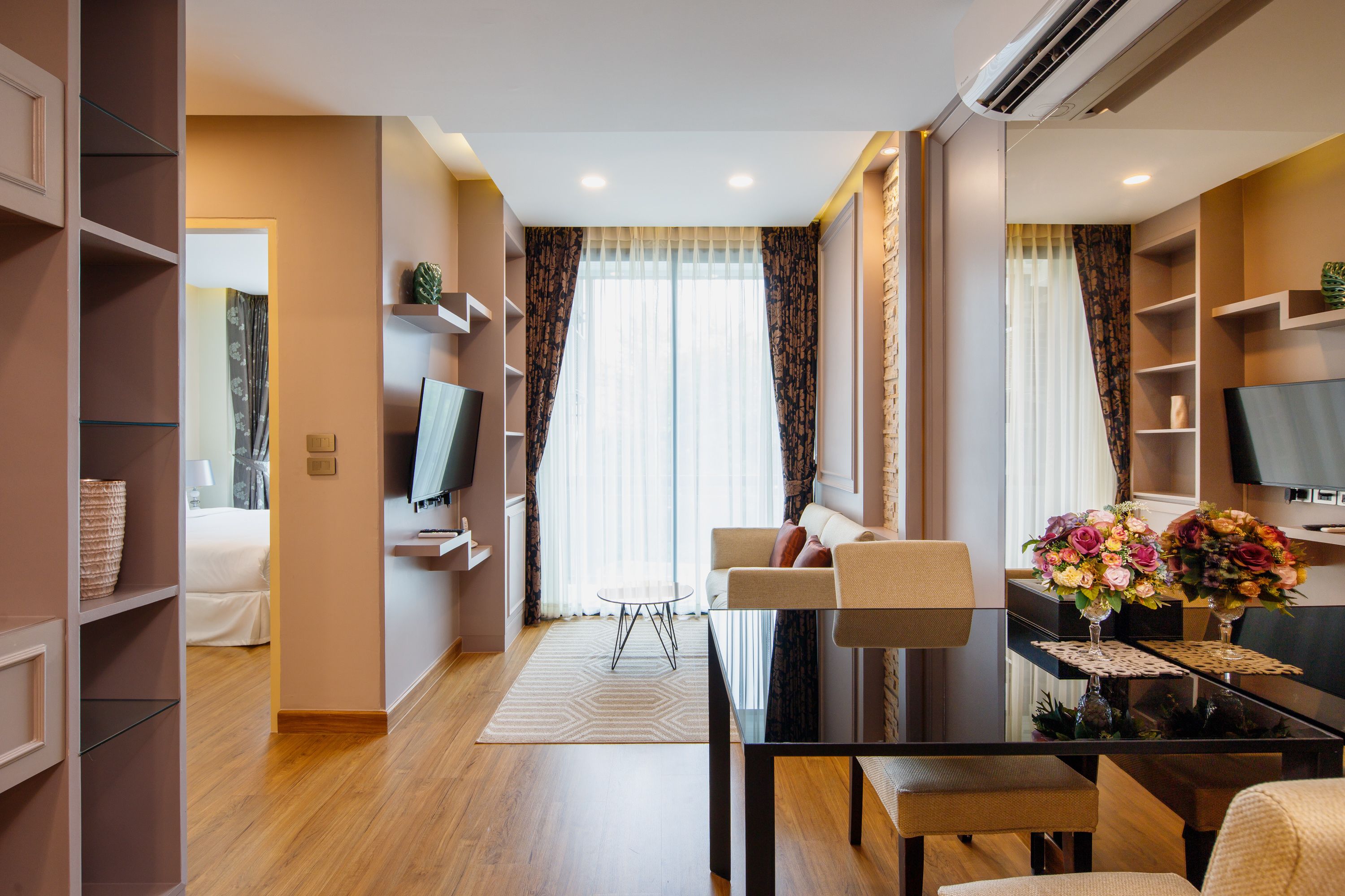 Una habitación de hotel moderna con zona de comedor, sofá, televisión y un amplio ventanal con cortinas.