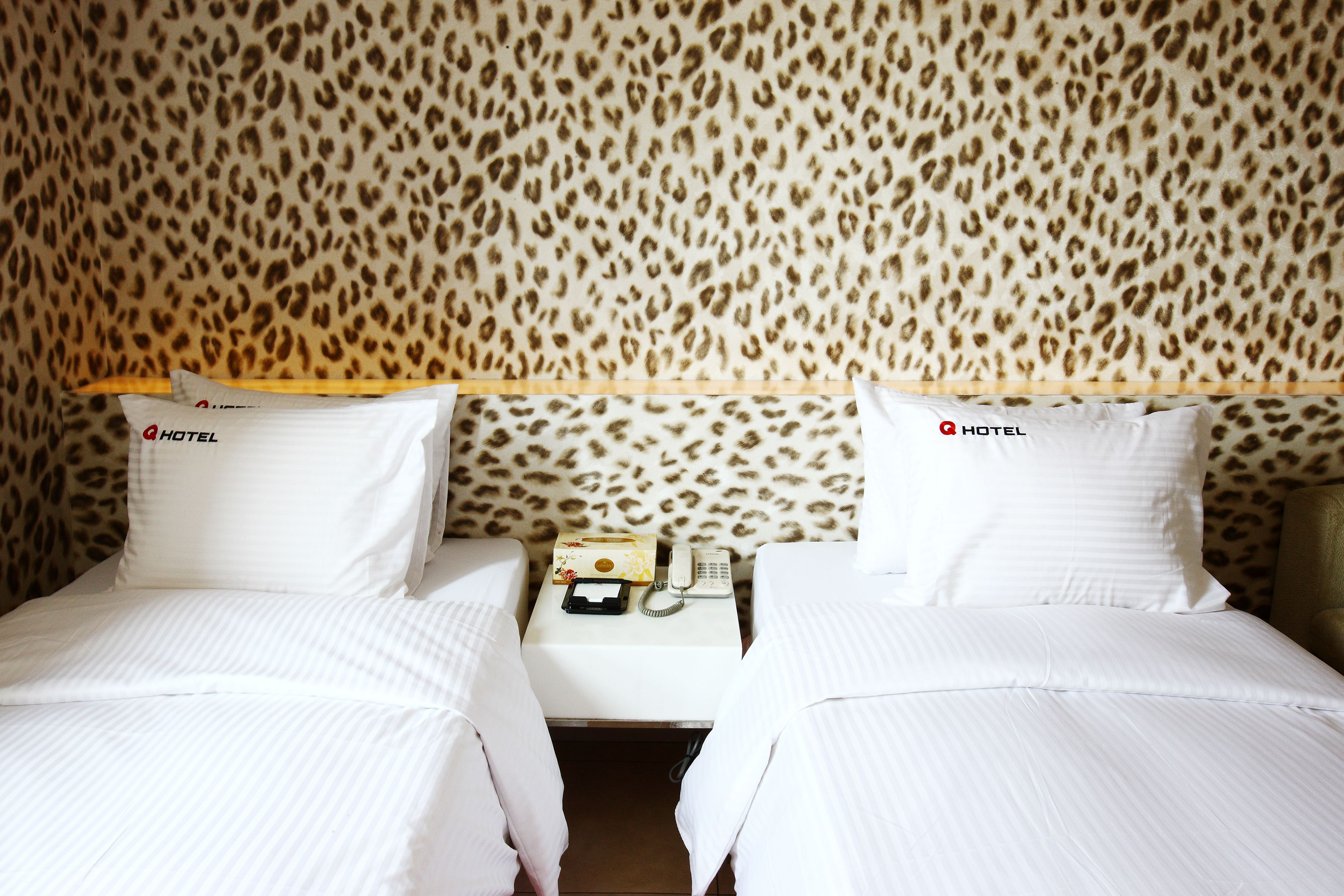 A hotel room with two beds, leopard print wallpaper, and a small table with a telephone and a box.