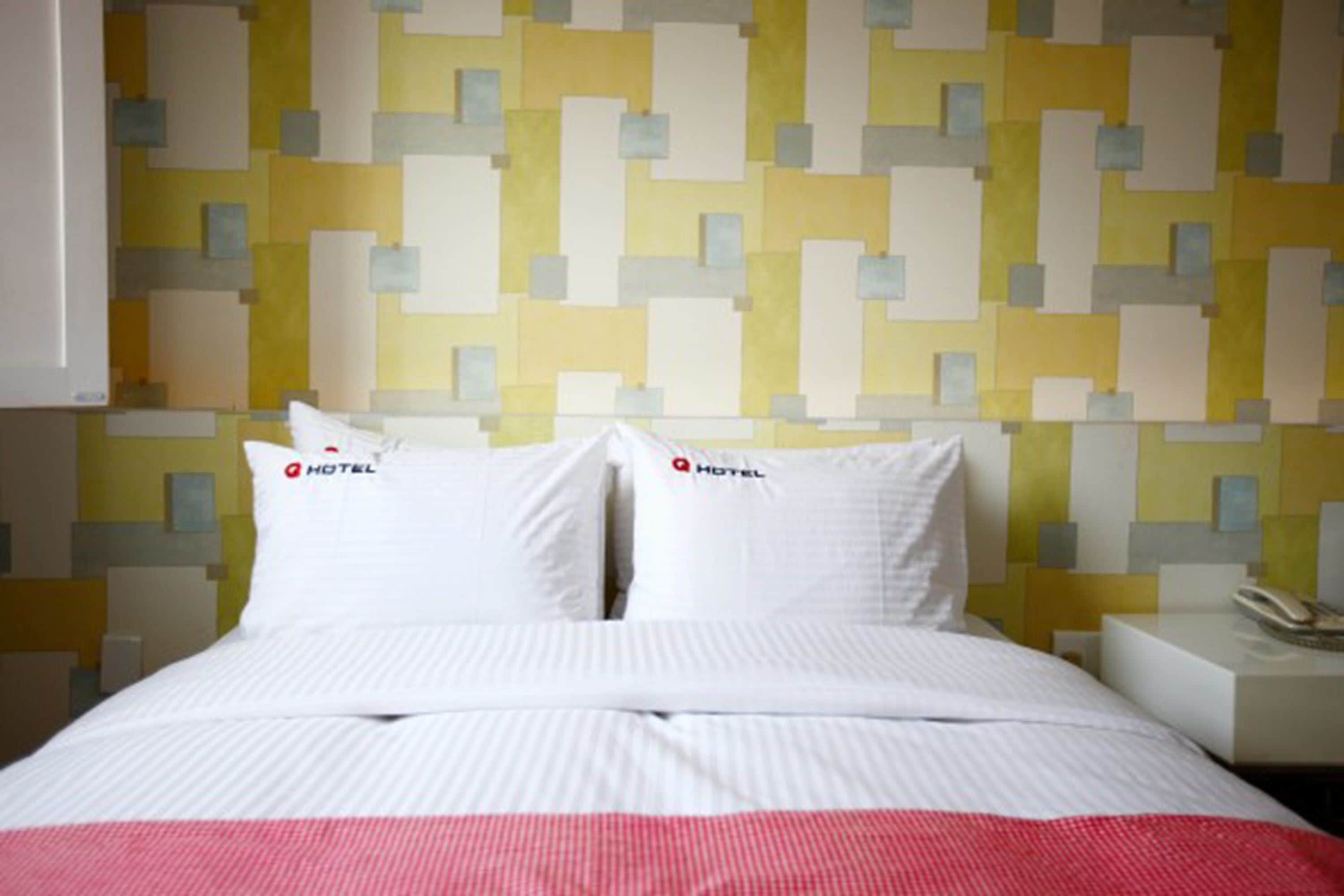 Una habitación de hotel con una cama, almohadas blancas con el logotipo del hotel, una mesita de noche con un teléfono y una pared con un diseño geométrico.