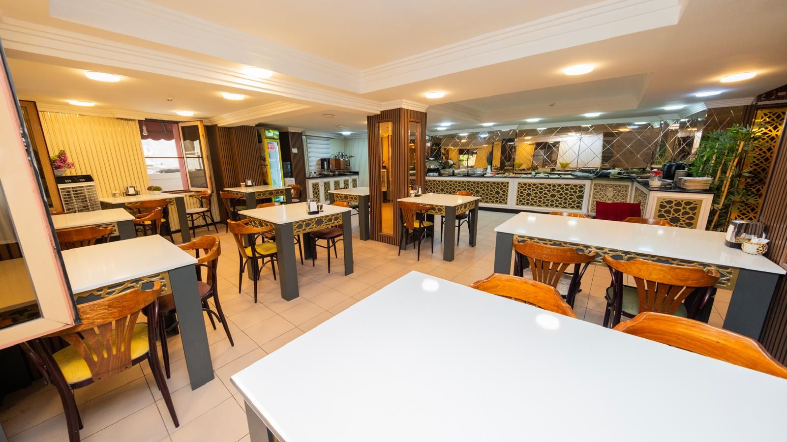 A restaurant with multiple dining tables and chairs, a kitchen area, and a decorative wall with geometric patterns.