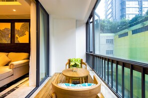 Un balcón con mesa y sillas de madera, un sofá y un ventanal con vistas a un edificio y áreas verdes.