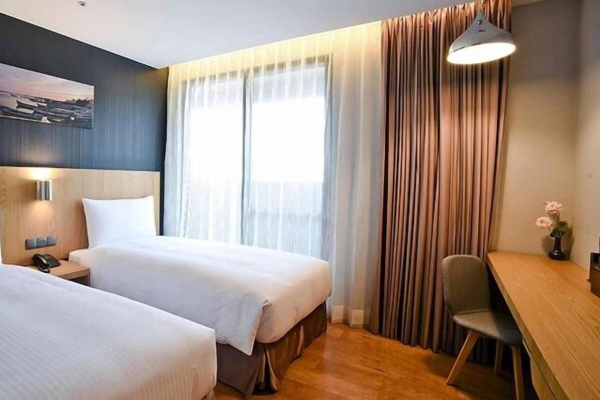 A hotel room with two beds, a desk, a chair, a lamp, and a painting on the wall.