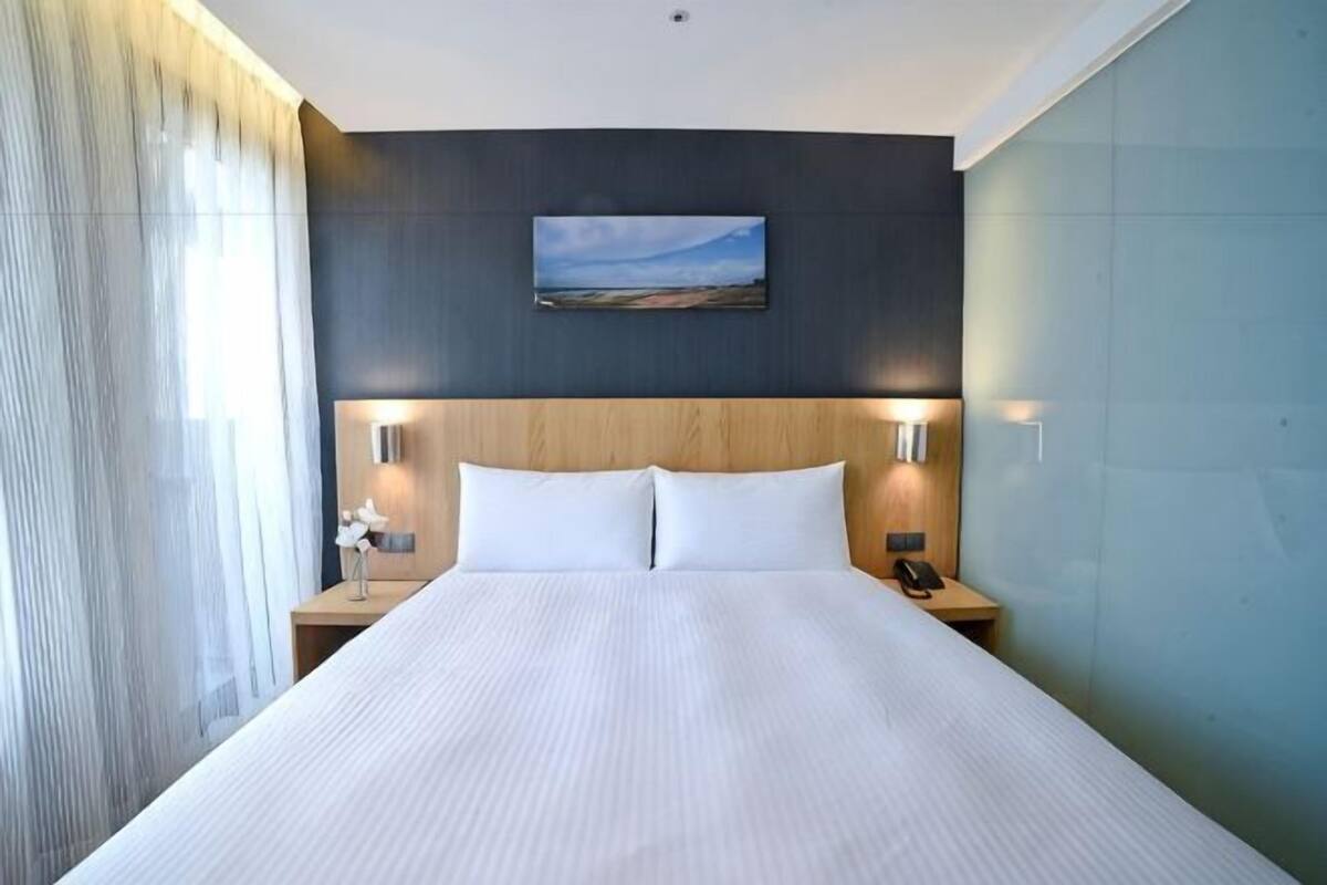 A hotel room with a large bed, two bedside tables, a nightstand, a painting on the wall, and a window with curtains.