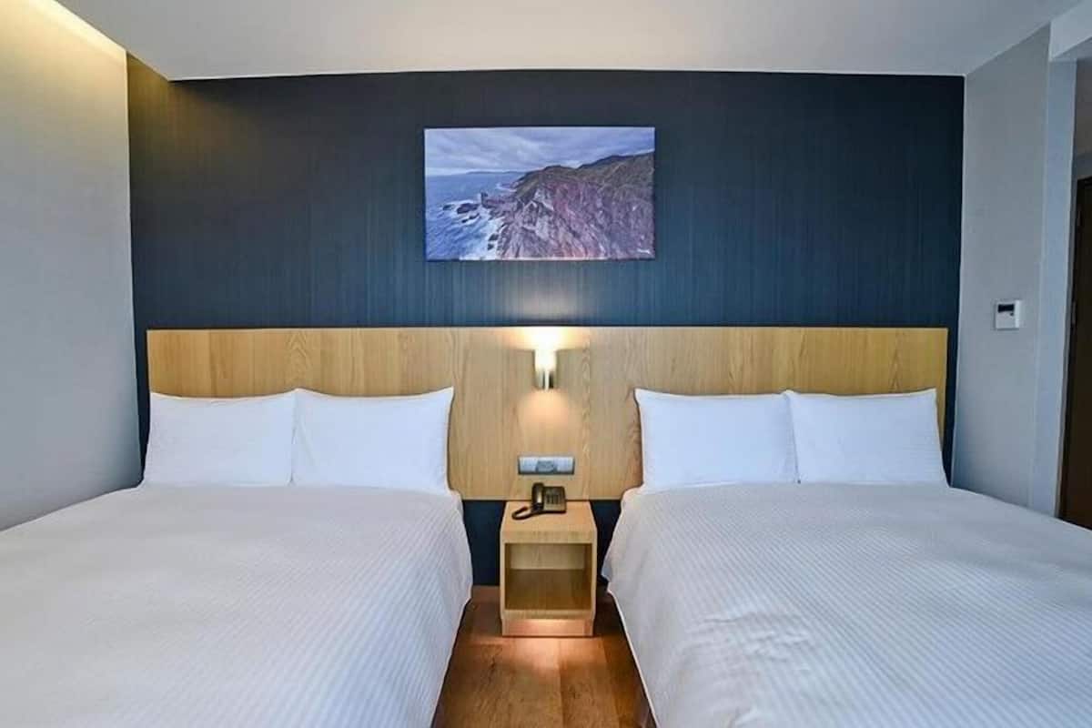 A hotel room with two beds, a nightstand with a phone, and a painting of a coastal landscape on the wall.