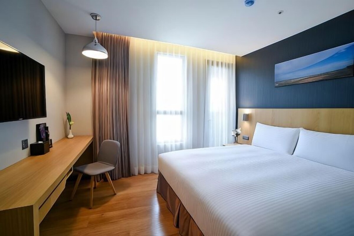 A hotel room with a large bed, a desk with a chair, a TV mounted on the wall, and a window with curtains.